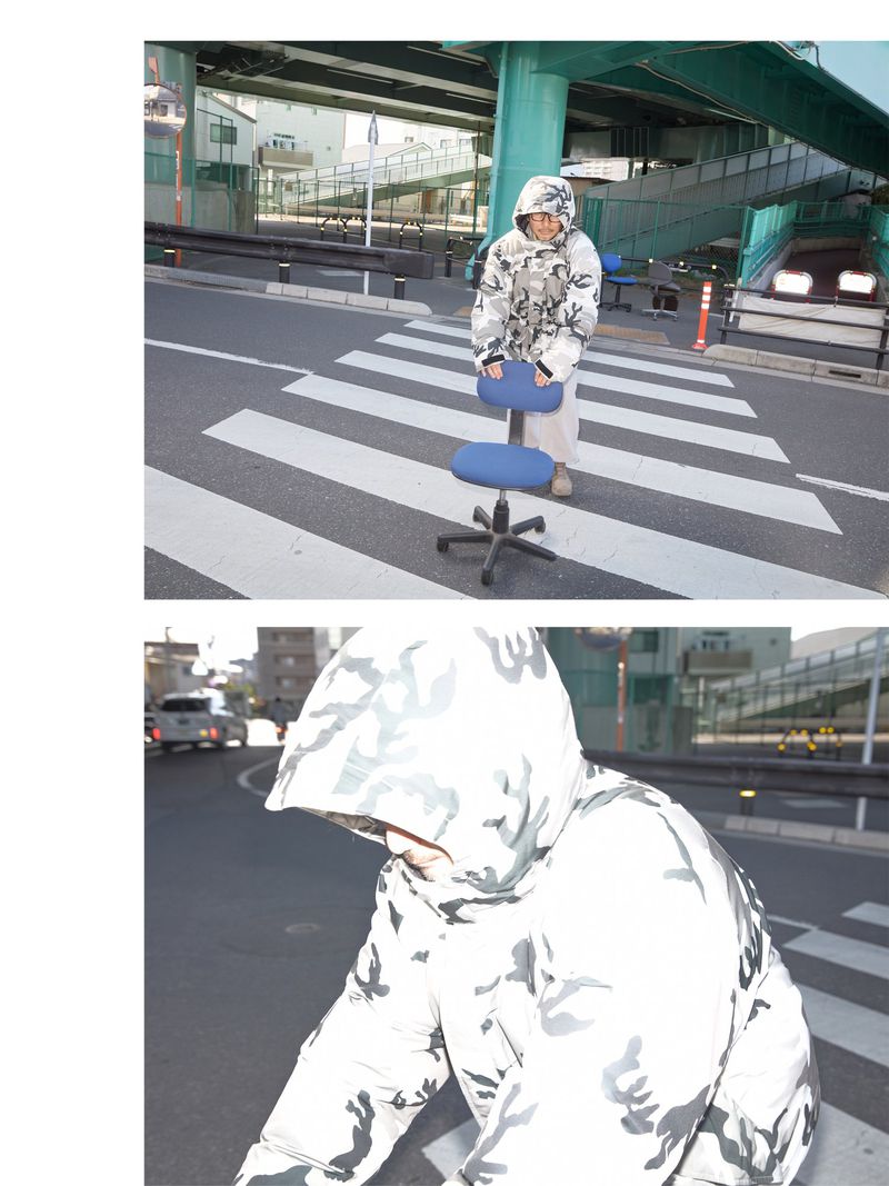 Adult wearing a camo hooded jacket sits on a rolling office chair in a city street crosswalk with overpass in the background.