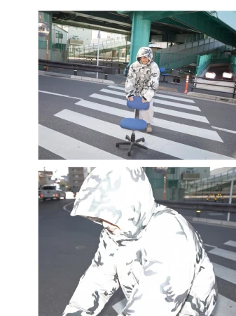 Adult wearing a camo hooded jacket sits on a rolling office chair in a city street crosswalk with overpass in the background.