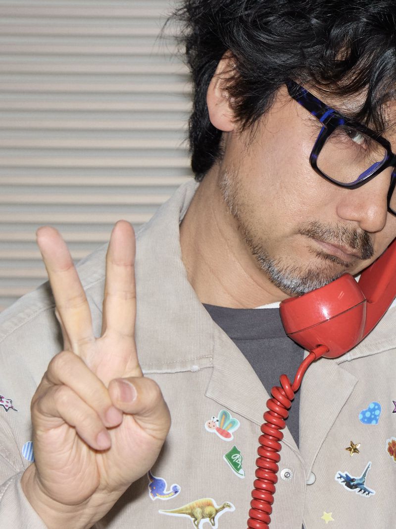 Adult man in glasses on a red phone, making a peace sign. He wears a light jacket with stickers. Indoor setting with window blinds.