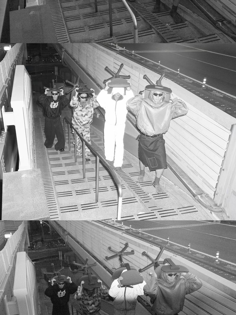Five people in costume walk down stairs, each carrying a chair on their heads. The image is black and white, creating a vintage effect.