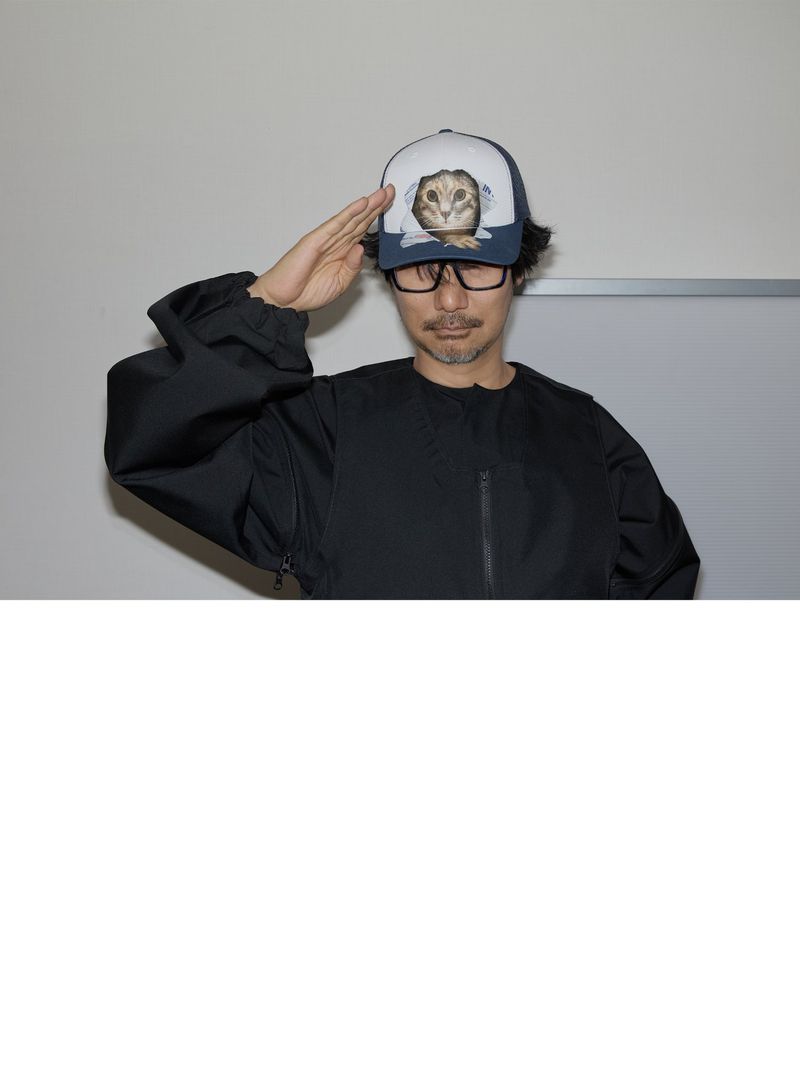 Man in a black jacket and cat-themed cap salutes indoors, against a plain wall.  Close-up, eye-level shot highlighting his eyewear and gesture.