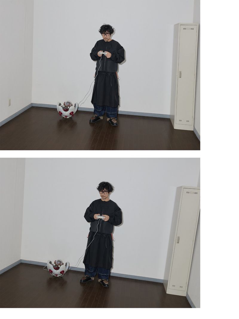 Adult wears glasses, a black jacket, and dark pants, playing a video game with a wired controller, indoors with a unique teapot-like object.