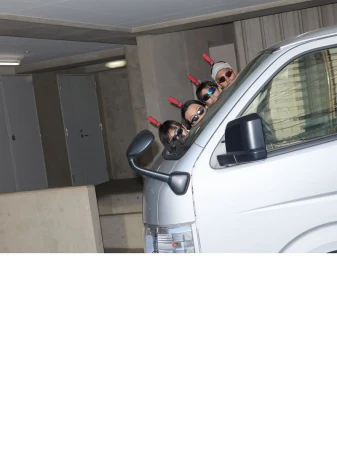 Four people wearing sunglasses and party antennas, leaning out of a silver van window with a cement wall in the background.