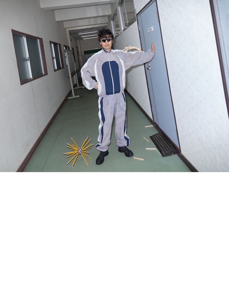 Man in a tracksuit posing in a hallway with pencils on the floor and a closed blue door next to him, wearing sunglasses.