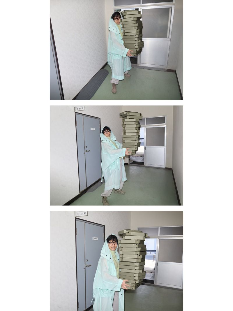 Person in light blue raincoat carries a tall stack of pizza boxes in a brightly lit hallway with doors on either side.