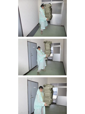 Person in light blue raincoat carries a tall stack of pizza boxes in a brightly lit hallway with doors on either side.