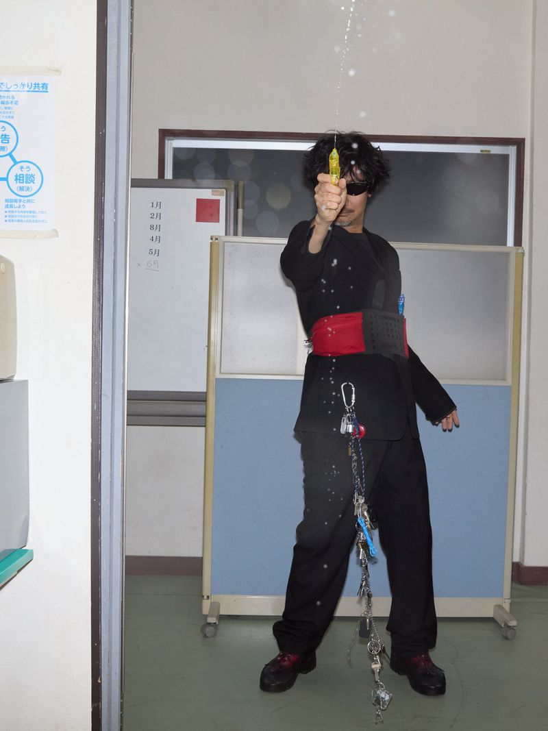 Person wearing black outfit stands indoors while spraying a yellow spray bottle; keychains hang from waist; indoor scene with light and white walls.