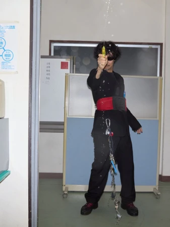 Person wearing black outfit stands indoors while spraying a yellow spray bottle; keychains hang from waist; indoor scene with light and white walls.
