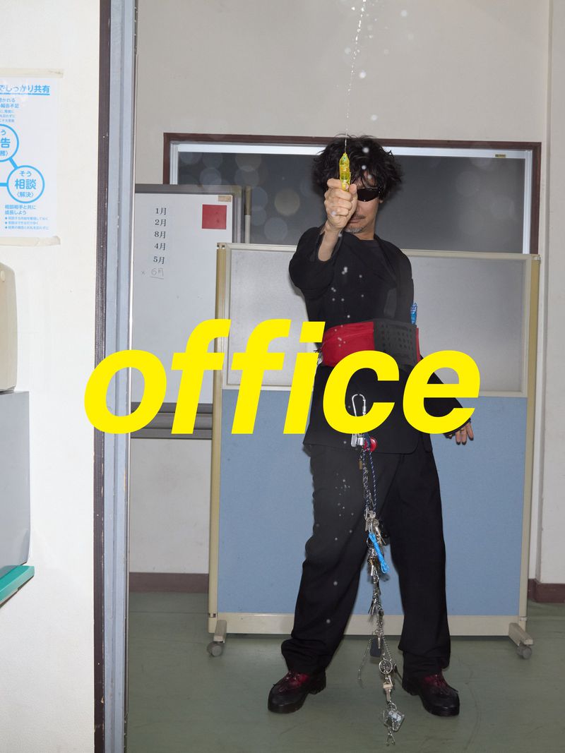 Man in black attire stands in an office, holding a yellow object with liquid spraying above, wearing sunglasses and a red belt. 