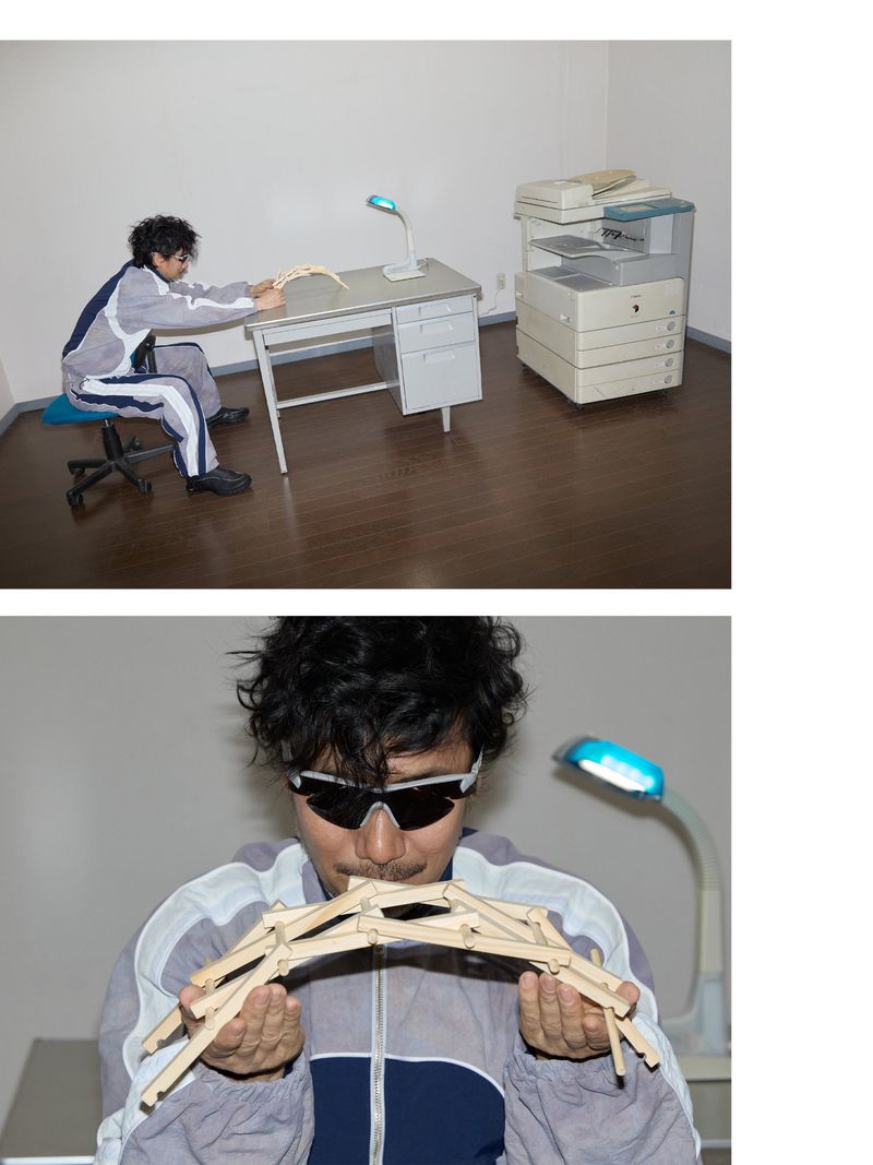Adult in grey tracksuit plays with wooden toy bridge at metal desk with a desk lamp in an office setting with a printer in the background.