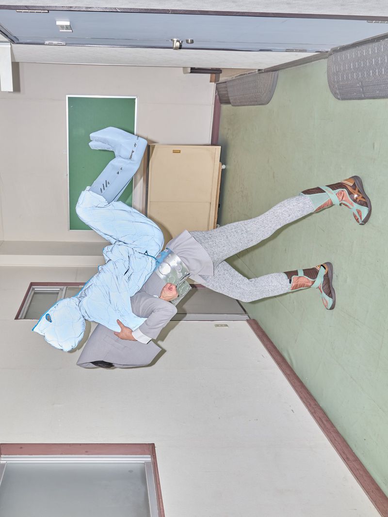 Two figures in abstract outfits pose against an indoor backdrop of walls, window, and door, creating a visually striking editorial image.