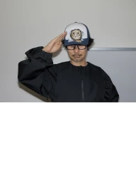 Man wearing black jacket and cat graphic cap saluting against plain wall