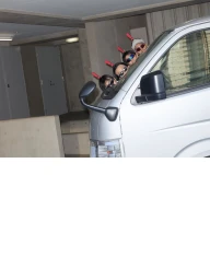 Four people wearing sunglasses and red accessories peek out from behind the front corner of a silver van in a parking garage.