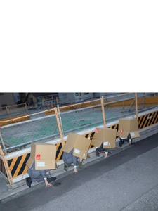 Four people crouching in a row on a street curb with cardboard boxes covering their heads and upper bodies beside a construction barrier.