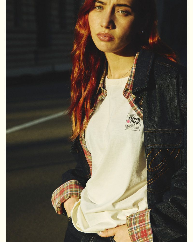 Young woman with red hair wearing a white t-shirt, plaid shirt, and dark denim jacket outdoors.