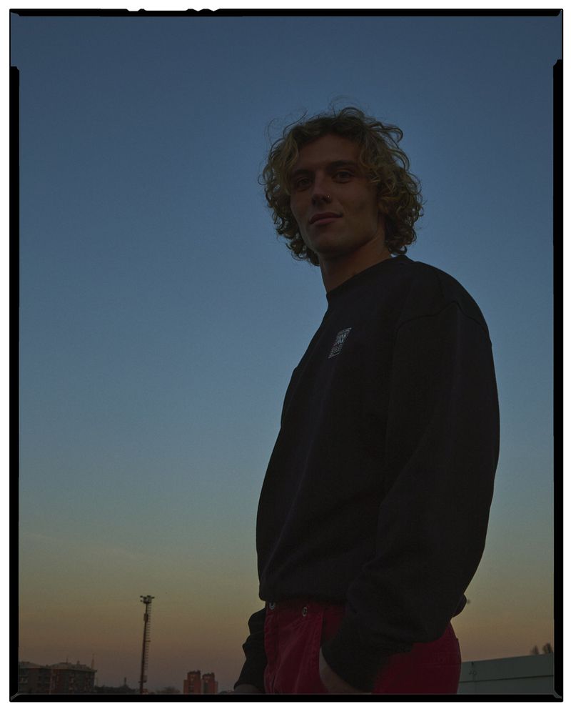 Young man with curly blonde hair wearing a dark sweatshirt and red trousers against a sunset sky.