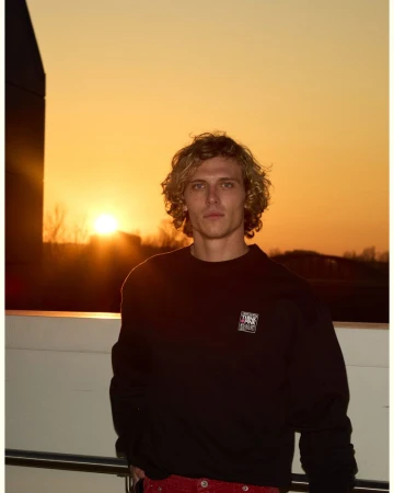 Young man with curly blonde hair wearing a black sweatshirt standing outdoors during a golden sunset.