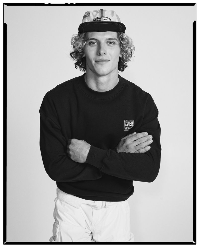 Black and white portrait of a curly-haired man wearing a patterned cap and dark sweatshirt with arms crossed.
