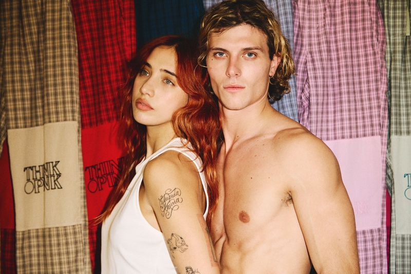 Close-up portrait of a young woman with reddish hair and a shirtless man standing closely against a colourful plaid textile backdrop.