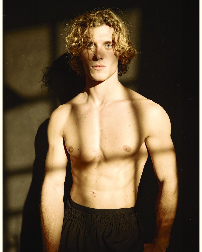 Shirtless young man with curly blonde hair posing against a dark wall under dramatic striped sunlight.