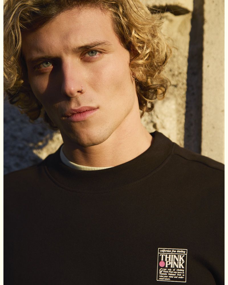 Close-up of a young adult male with curly blonde hair wearing a black sweatshirt and posing in an outdoor setting with textured walls.
