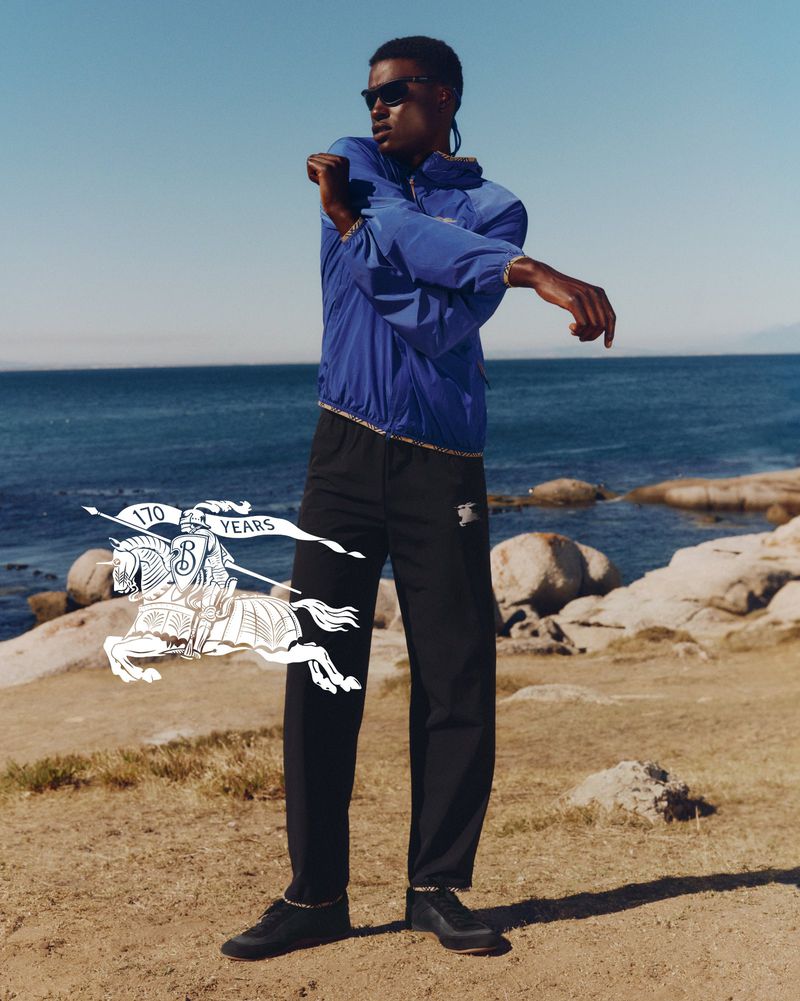 Model wearing a blue jacket and black trousers stretching by a rocky coast against the ocean and sky