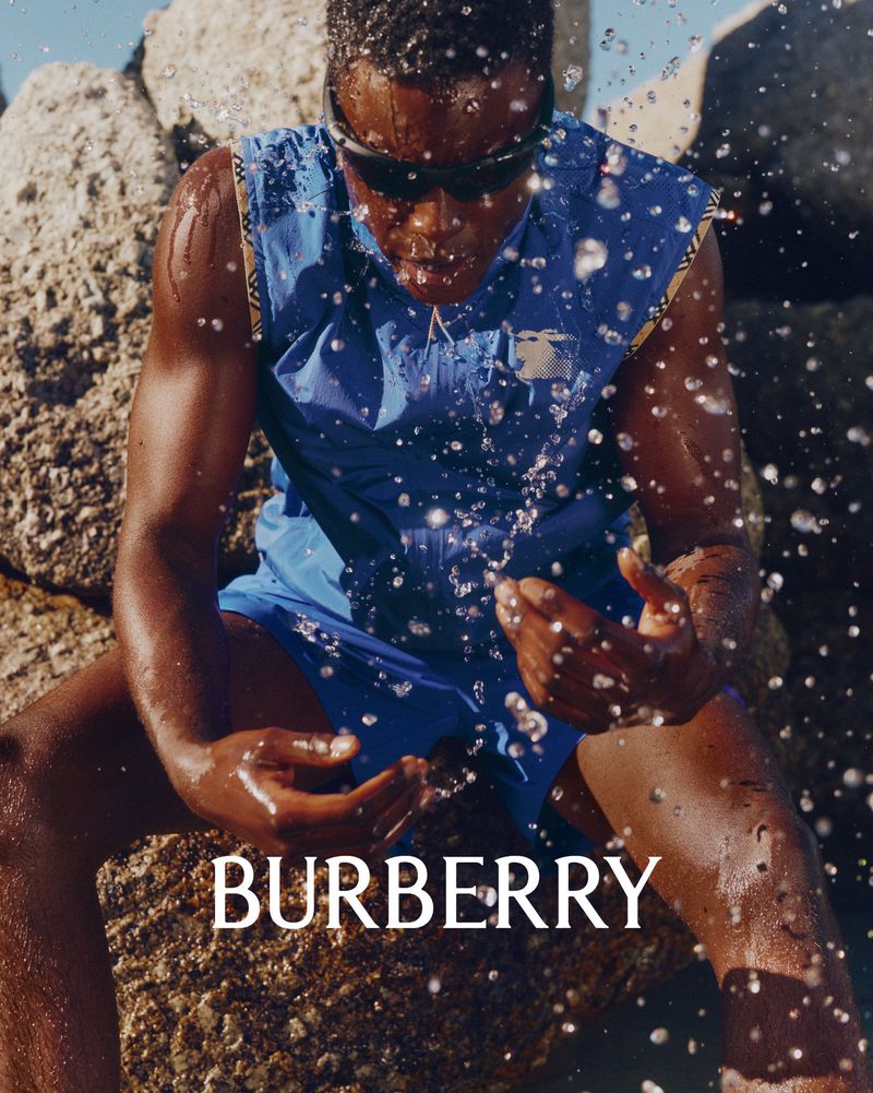 Model wearing blue athletic vest and sunglasses sitting among rocky boulders with splashing water drops