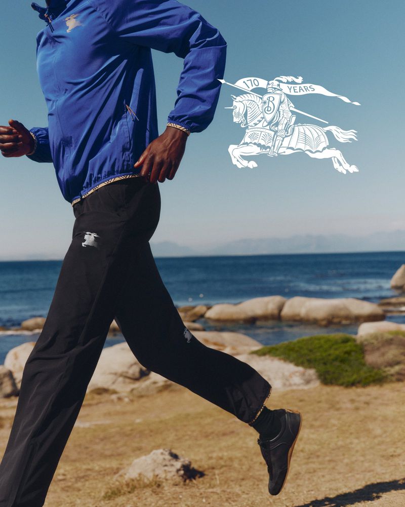 Runner in blue jacket and black trousers jogging along a coastal path with a white graphic knight emblem in the sky.