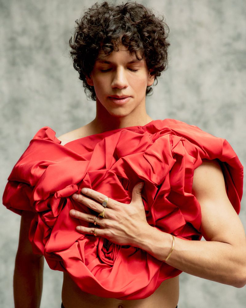 Adult male with curly hair and a red fabric covering his chest, posing against a gray backdrop in a portrait-style studio shot.