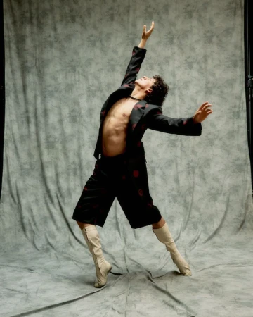 Adult male dances in a black blazer and shorts with red rose patterns in front of a gray mottled background in a photo studio.