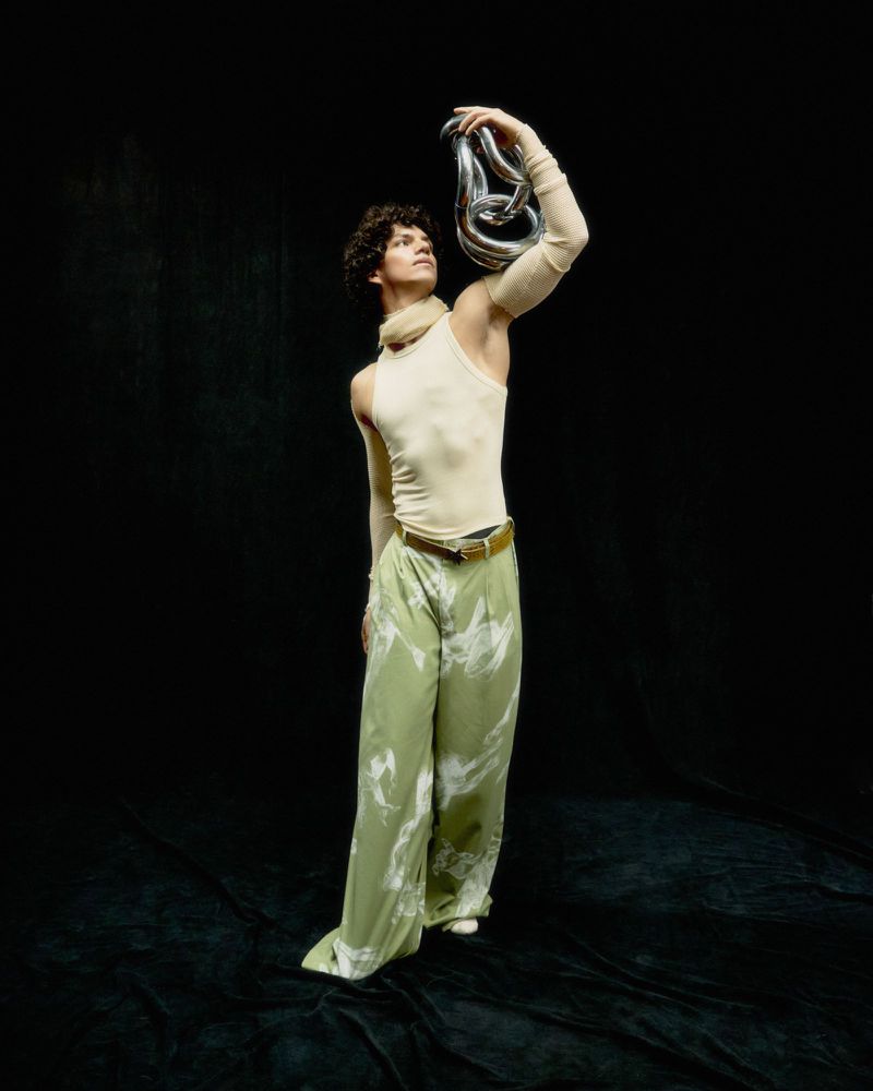 Adult male model wearing an ivory tank top and green patterned pants poses with linked silver chain against a dark backdrop.