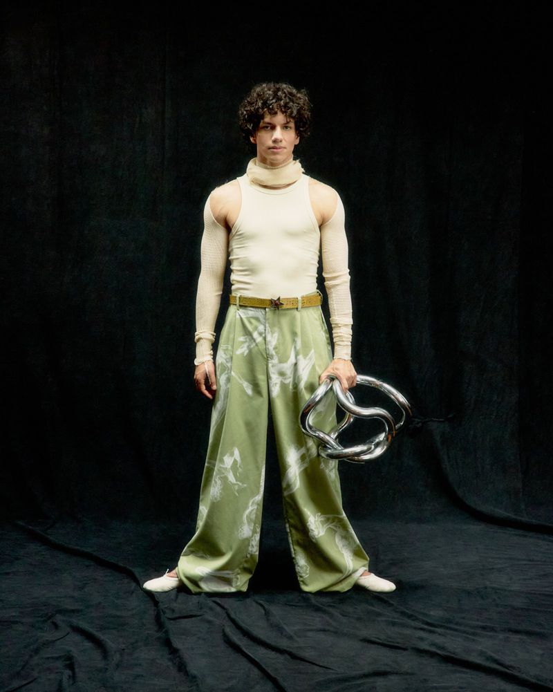 A young adult male with curly hair poses in an indoor studio wearing a turtleneck, patterned pants, and holding a twisted metal prop.