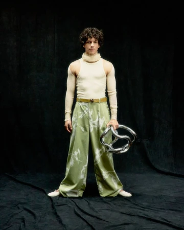 A young adult male with curly hair poses in an indoor studio wearing a turtleneck, patterned pants, and holding a twisted metal prop.
