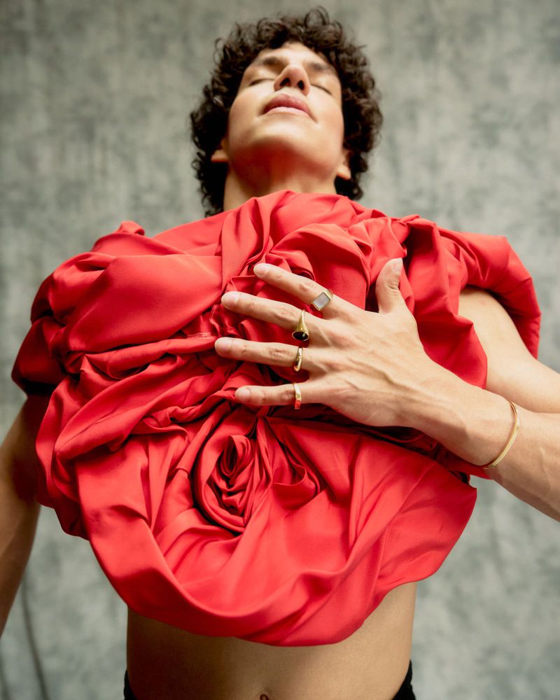 A person with curly hair poses indoors wearing a large, red, floral-shaped garment, showcasing its intricate folds and design in a fashion-forward shot.