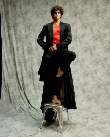 Ballet dancer Isaac Hernández posing with one foot raised on a metal stool against a mottled grey textured studio backdrop.