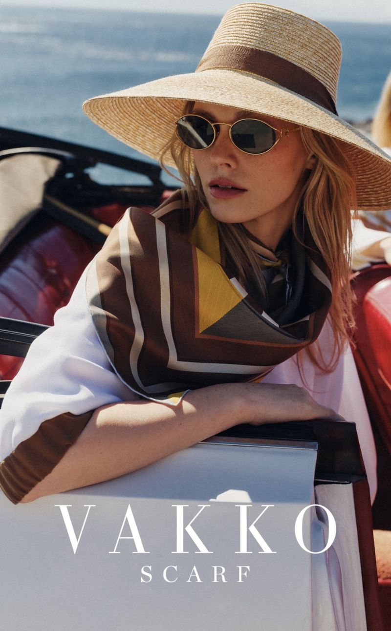 Model wearing a straw sun hat, sunglasses, and a patterned silk scarf while leaning against a red car with the ocean behind