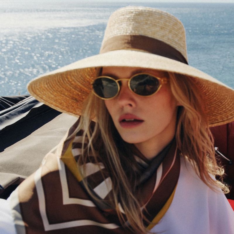 Blonde woman in sunglasses and a large straw hat with a brown band sits outdoors on a boat with the ocean in the background.
