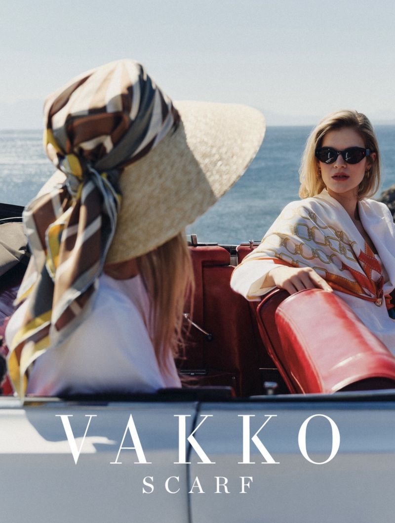 Two women in a convertible car near the ocean, wearing printed silk scarves and sunglasses under bright sunlight