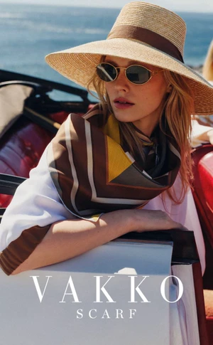Model wearing a straw sun hat, sunglasses, and a patterned silk scarf while leaning against a red car with the ocean behind