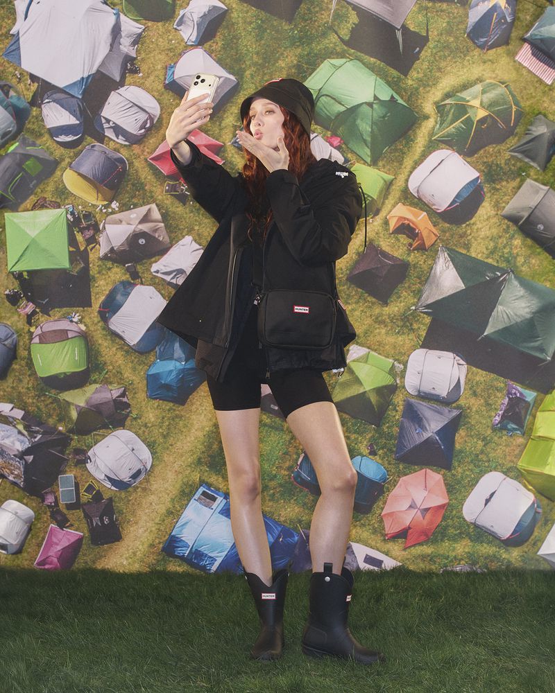 Model wearing black boots and jacket taking a selfie against a backdrop of colorful festival tents