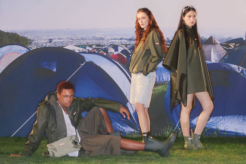 Three models in green raincoats and rubber boots posing outdoors against a background of festival tents.