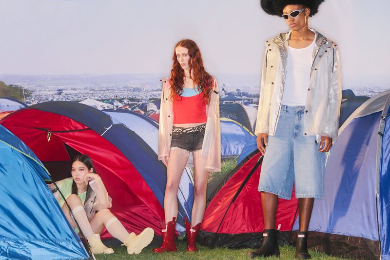 Three fashion models in rain gear stand in front of colorful tents, evoking a festival atmosphere on an overcast day.