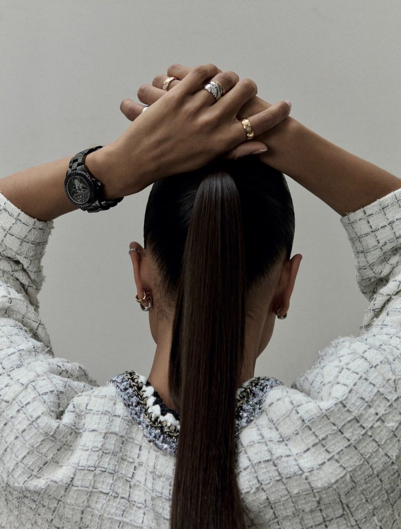 Back view of a woman with a sleek ponytail wearing a patterned jacket, showcasing a watch and rings, against a plain background.
