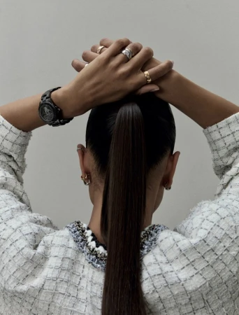 Back view of a woman with a sleek ponytail wearing a patterned jacket, showcasing a watch and rings, against a plain background.