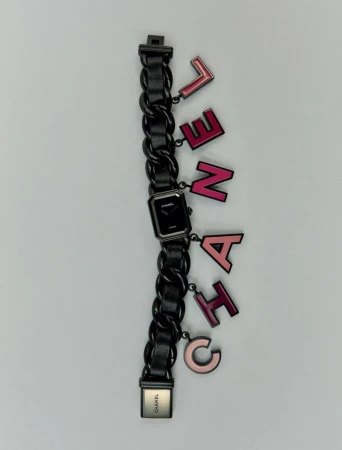 Chanel Première watch with black chain-link bracelet and letter charms, studio shot against a light gray background.