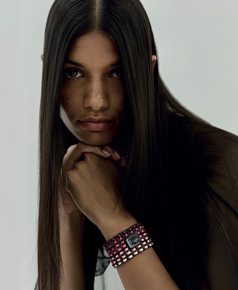 Close-up of a model with long, straight brown hair with a colorful woven wristwatch posing in front of a neutral background.