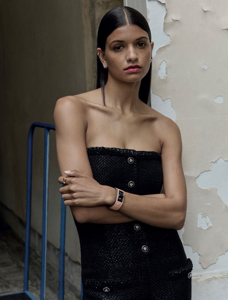 Woman in strapless black dress with watch poses near weathered wall, showcasing fashion and portrait photography in a neutral indoor setting.