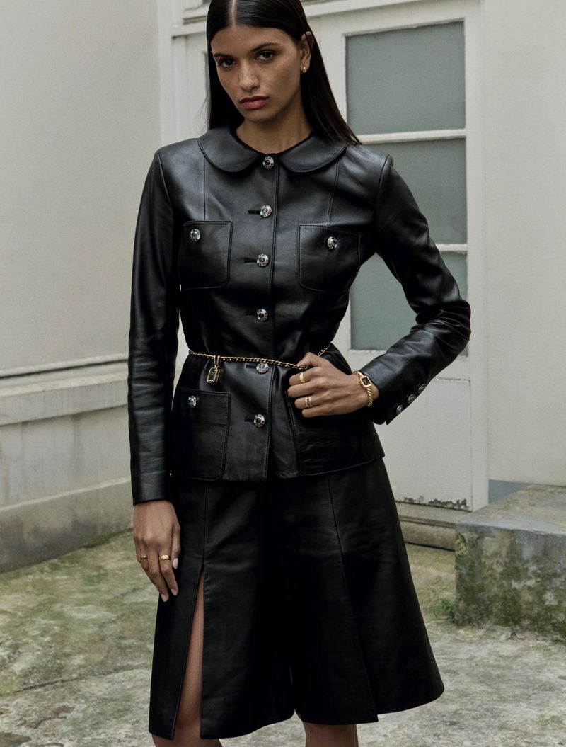 Brunette woman in a black leather jacket and skirt, accessorized with gold jewelry, posing outdoors against a neutral-toned building.