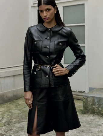 Brunette woman in a black leather jacket and skirt, accessorized with gold jewelry, posing outdoors against a neutral-toned building.