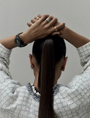 Back view of a woman with a sleek high ponytail and hands behind her head wearing a white tweed jacket, stacked rings, and a black watch.